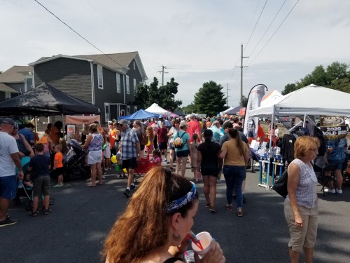 A busy street fair with crowds of people, tents, and vendors under a sunny sky.