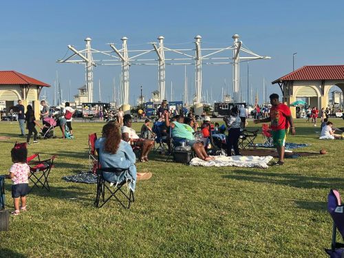 A lively outdoor gathering at a marina, with families sitting on blankets and chairs, enjoying the sunny day.