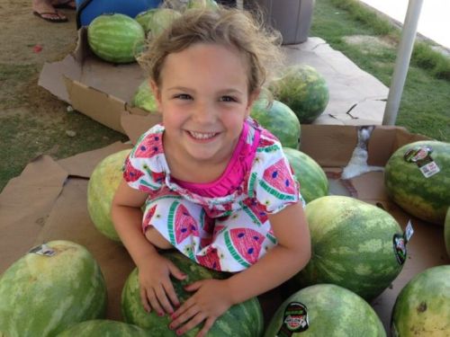 A smiling girl sits among watermelons, wearing a colorful watermelon-patterned dress, on a sunny day.