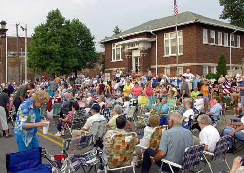 A large crowd of people seated in lawn chairs outside a library, enjoying a community event on a sunny day.