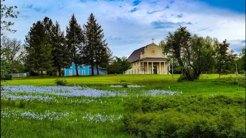 A serene landscape featuring a church, vibrant green grass, and a field of blue flowers under a cloudy sky.