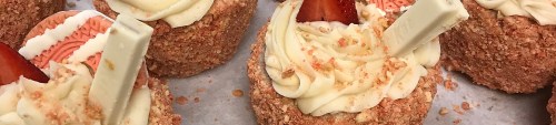 Close-up of cupcakes topped with creamy frosting, strawberry slices, and white chocolate sticks, sprinkled with crumbs.