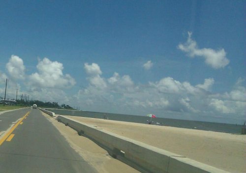 A scenic view of a beachside road with a sandy shore, colorful umbrellas, and a blue sky dotted with clouds.
