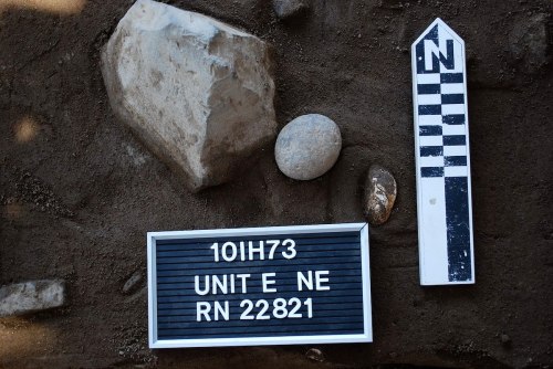 A sign with "10IH73 UNIT E NE RN 22821" next to rocks and a north marker on a sandy surface.