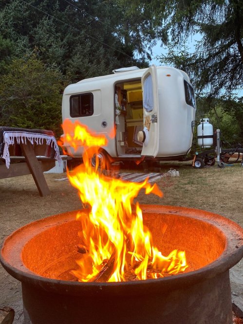 A cozy campsite with a fire pit in the foreground and a small camper trailer in the background, surrounded by trees.