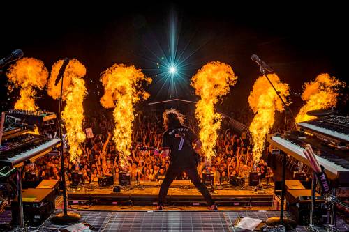 A guitarist performs on stage with flames shooting up behind him, while a cheering crowd enjoys the show.