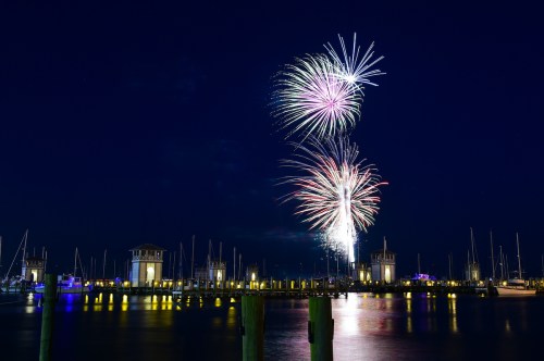 Colorful fireworks burst over a marina at night, reflecting on the water with boats and illuminated buildings in the background.