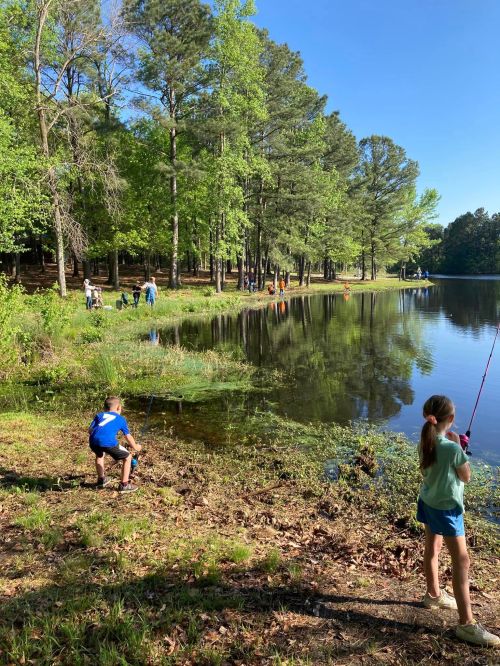 Children fishing by a serene pond surrounded by trees, with others enjoying the outdoors in the background.