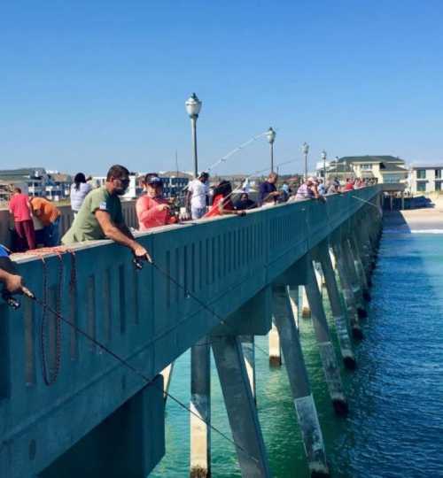 People fishing from a pier on a sunny day, with clear blue water and a bright sky in the background.