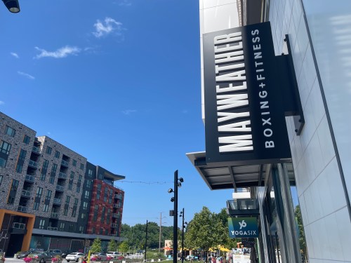 Sign for Mayweather Boxing + Fitness under a clear blue sky, with modern buildings and a Yogasix sign in the background.