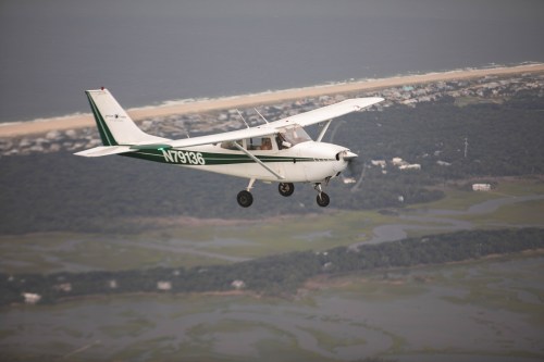 A small airplane flying over a coastal landscape with water and greenery below.