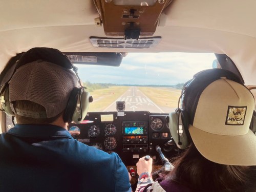 Two pilots in a small aircraft, wearing headsets, preparing for takeoff on a runway with a clear sky ahead.
