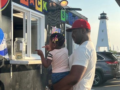A man holds a girl as she points at a food truck window, with a lighthouse in the background.