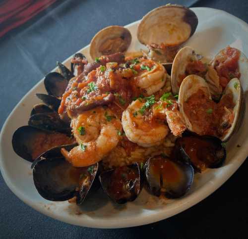 A plate of seafood featuring shrimp, clams, and mussels served over rice with a rich tomato sauce and herbs.