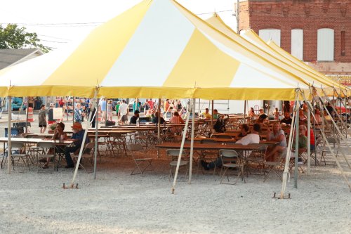 A large yellow and white tent with tables and chairs, set up in a gravel area, with people gathered around.