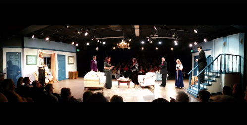 A theater stage with actors in formal attire performing a scene, surrounded by an audience in a dimly lit venue.