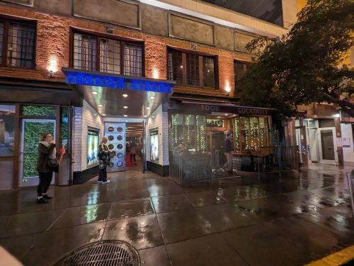 A lively street scene at night featuring a cinema and a restaurant with outdoor seating, illuminated by warm lights.