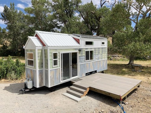 A rustic tiny house on wheels with a wooden deck, surrounded by trees and open land under a blue sky.