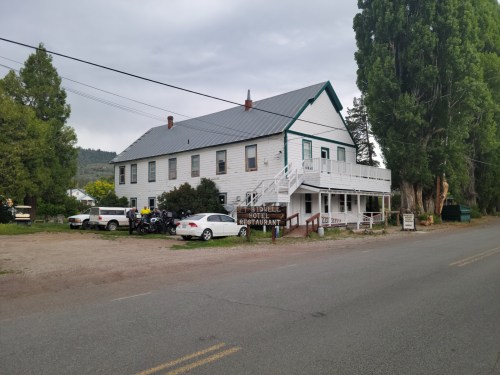A two-story white building with a gray roof, featuring a restaurant sign, parked cars, and trees nearby.