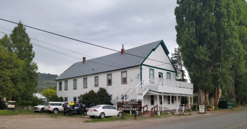 A two-story white building with a gray roof, featuring a porch and sign for a hotel and restaurant, surrounded by trees.