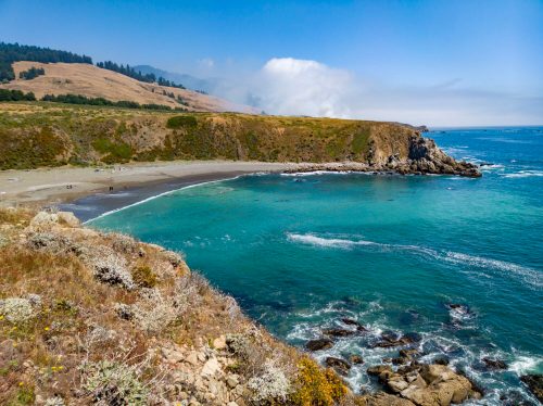 A scenic coastal view featuring a sandy beach, rocky shoreline, and clear blue waters under a bright sky.