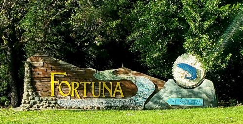 Sign for Fortuna featuring colorful lettering and a fish design, surrounded by greenery.