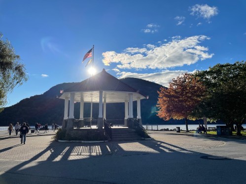 A gazebo with a flag stands against a backdrop of mountains and a bright sky, with people walking nearby.