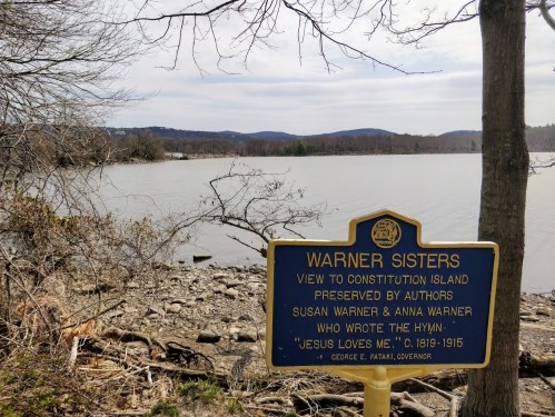 A sign about the Warner Sisters near a calm lake, with trees and mountains in the background under a cloudy sky.