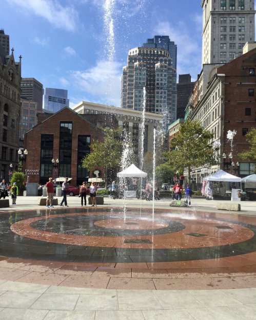A fountain in a city square, surrounded by buildings, trees, and people enjoying the sunny day.
