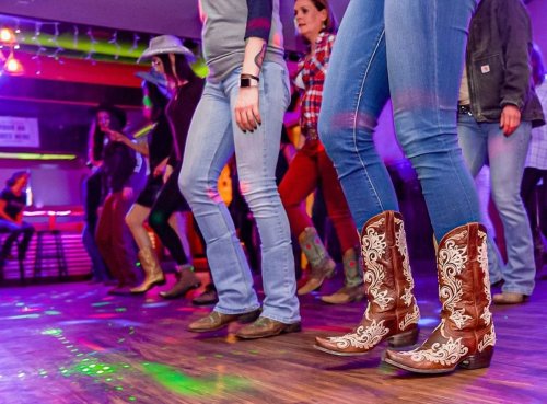 A group of people line dancing in cowboy boots, with colorful lights reflecting on the dance floor.