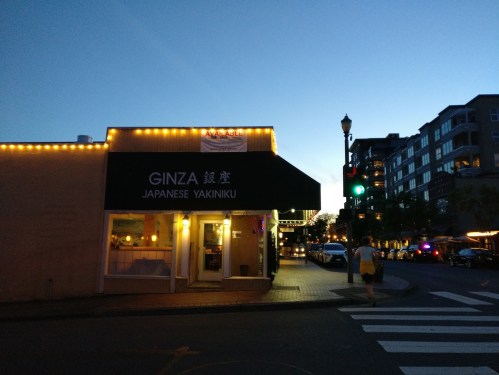 A street view of Ginza Japanese Yakiniku restaurant at dusk, with city buildings and traffic lights in the background.