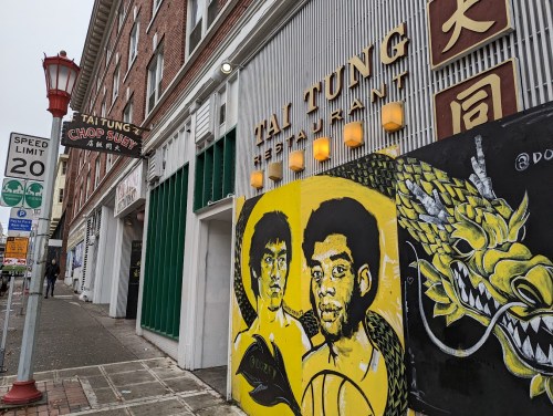 A street view of Tai Tung restaurant with a colorful mural featuring two basketball players on the wall.