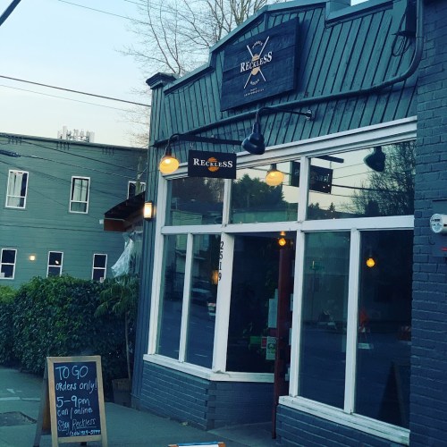 Exterior of a restaurant named "Reckless," featuring large windows and a sign, with a green hedge in front.