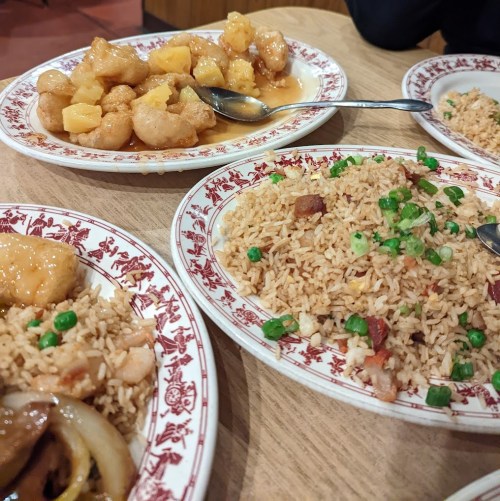 Two plates of fried rice with green onions and a plate of sweet and sour shrimp with pineapple on a wooden table.