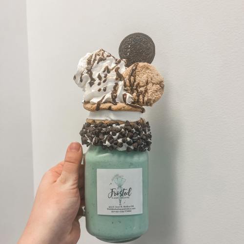 A hand holding a tall milkshake topped with whipped cream, a cookie, an Oreo, and chocolate drizzle.