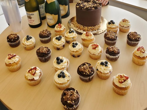 A table with a chocolate cake and assorted cupcakes topped with fruit and nuts, surrounded by bottles of champagne.