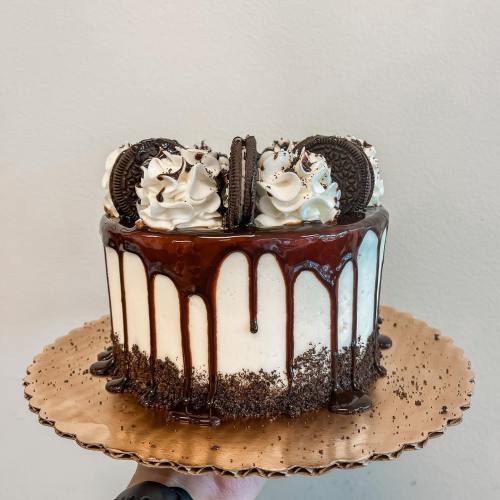 A round cake topped with whipped cream, Oreos, and chocolate drizzle, sitting on a gold plate.