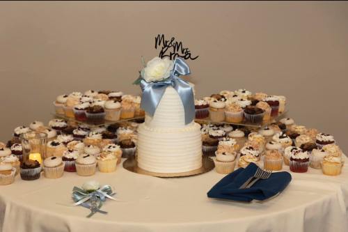 A tiered wedding cake with a floral decoration surrounded by an array of assorted cupcakes on a table.