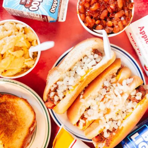 A colorful spread of two hot dogs topped with onions, macaroni and cheese, baked beans, and a slice of toast.