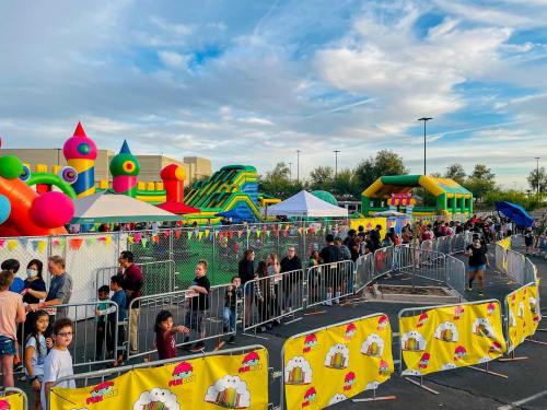 A lively outdoor event with colorful bounce houses and a long line of people waiting, surrounded by festive decorations.