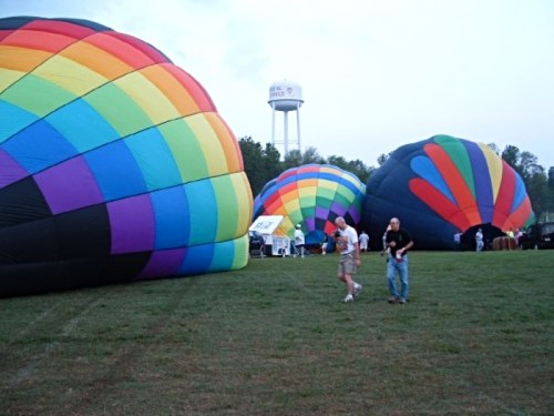 Colorful hot air balloons are being inflated on a grassy field, with a water tower visible in the background.