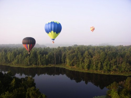 Three hot air balloons float over a serene lake surrounded by lush green trees in the early morning mist.
