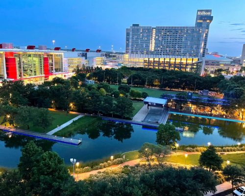 A scenic view of a park and waterway at dusk, with a Hilton hotel and city buildings in the background.