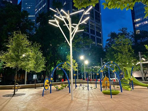 A modern playground at night, featuring colorful equipment and illuminated trees, surrounded by tall buildings.