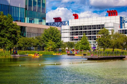 A serene park scene with kayakers on a calm lake, surrounded by greenery and modern buildings under a blue sky.