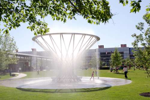 A large, modern fountain in a sunny park, with people enjoying the greenery and water features around it.