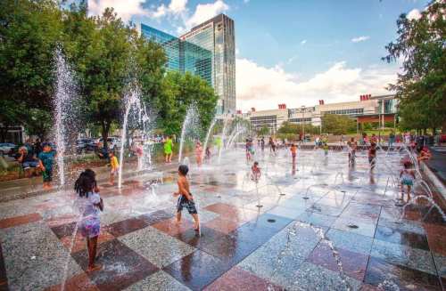 Children play in a splash pad, surrounded by fountains, with buildings and trees in the background on a sunny day.