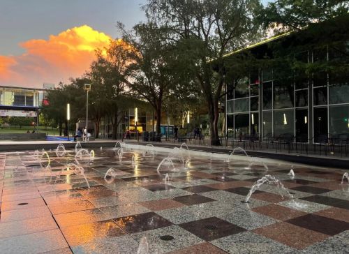 A plaza with water fountains, surrounded by trees and modern buildings, under a colorful sunset sky.