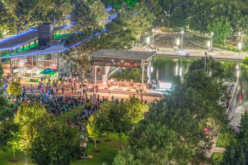 Aerial view of a lively outdoor concert by a lake, with a crowd enjoying the performance amidst trees and lights.