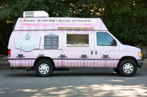 A pink food truck with "Cupcake City" branding, featuring a menu of cupcake flavors and contact information.